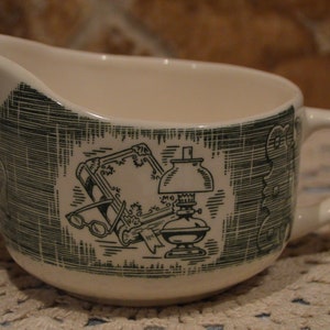 May include: Cream-colored gravy boat with a green and white illustration. The illustration depicts a lamp, glasses, and a book. The gravy boat has a handle and a spout.