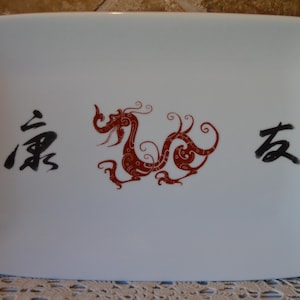 May include: White rectangular serving platter with a red dragon design in the center. The dragon is surrounded by black Chinese characters. The platter has rounded corners and a glossy finish.