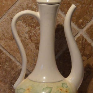 May include: A white ceramic jug with a floral design. The jug has a tall, slender neck and a wide, round base. The floral design features yellow flowers and green leaves.
