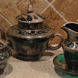 May include: A silver and green tea set with a teapot, creamer, and sugar bowl. The set has an ornate design with silver accents on a green background.