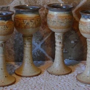 Può includere: Quattro calici in ceramica fatti a mano con finitura beige e marrone macchiettata. Ogni calice ha una coppa arrotondata, un lungo stelo e una base larga. I bordi sono blu scuro. I calici sono esposti su una tovaglia di pizzo bianco.