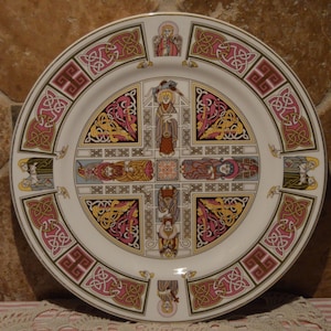 May include: A white ceramic plate with a Celtic knot design in gold, red, and green. The center of the plate features a cross with figures in the four quadrants.