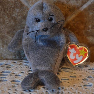 May include: A grey plush seal toy with black eyes and whiskers. The seal has a tag attached with a red heart shape and the words "Ty Beanie Baby". The toy is sitting on a white lace doily.