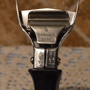 May include: Close-up of a vintage Corning Ware can opener. The can opener is made of silver metal with a black handle. The text "CORNING WARE" is visible on the metal. The can opener has a classic design.