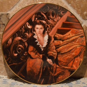 May include: A decorative plate featuring a portrait of a woman in a period costume. The plate has a gold rim and a detailed illustration of a woman in a fur-trimmed coat and a ruffled collar, seated near a staircase.