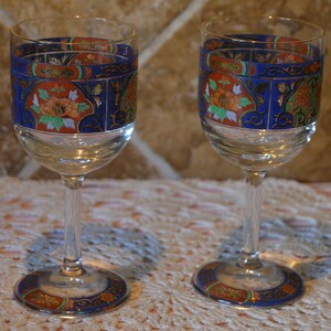Op de afbeelding: Twee heldere wijnglazen met sierlijke blauwe, oranje en gouden bloemmotieven. De glazen hebben een heldere steel en voet, met hetzelfde decoratieve patroon op de voet als op de rand. Neutrale achtergrond.