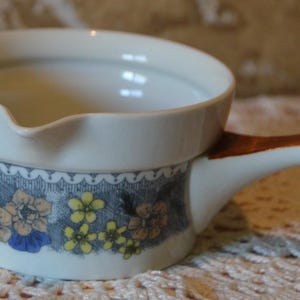 May include: A white ceramic soup bowl with a brown handle and a floral design. The bowl has a spout and a decorative band with blue, yellow, and orange flowers. The handle is curved and painted brown.