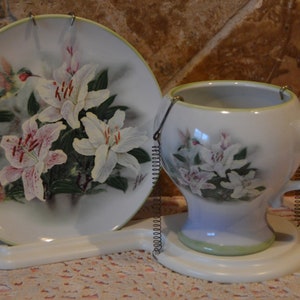 May include: A decorative teacup and saucer set. The white porcelain pieces feature a floral design of white and pink lilies with green leaves. A hummingbird is depicted on the plate. The set is connected by a metal spring.