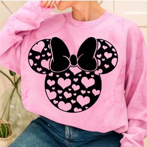 May include: Pink sweatshirt featuring a black Minnie Mouse design. The design includes a large bow and mouse ears filled with pink hearts. The sweatshirt has a tie-dye effect and is made of a soft material.