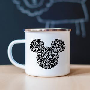 May include: White enamel mug with a silver rim and handle. The mug features a black, decorative Mickey Mouse head design. The design is a detailed mandala pattern. The mug is sitting on a light brown surface.