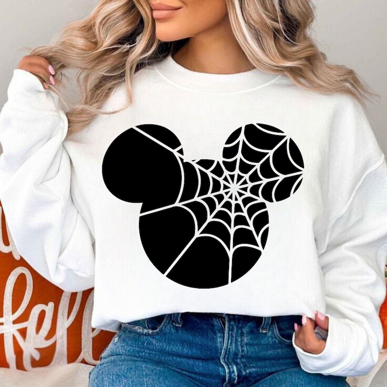 May include: White sweatshirt featuring a black Mickey Mouse-inspired design with a spiderweb. The graphic is centered on the front of the long-sleeved top. The sweatshirt is paired with blue jeans.
