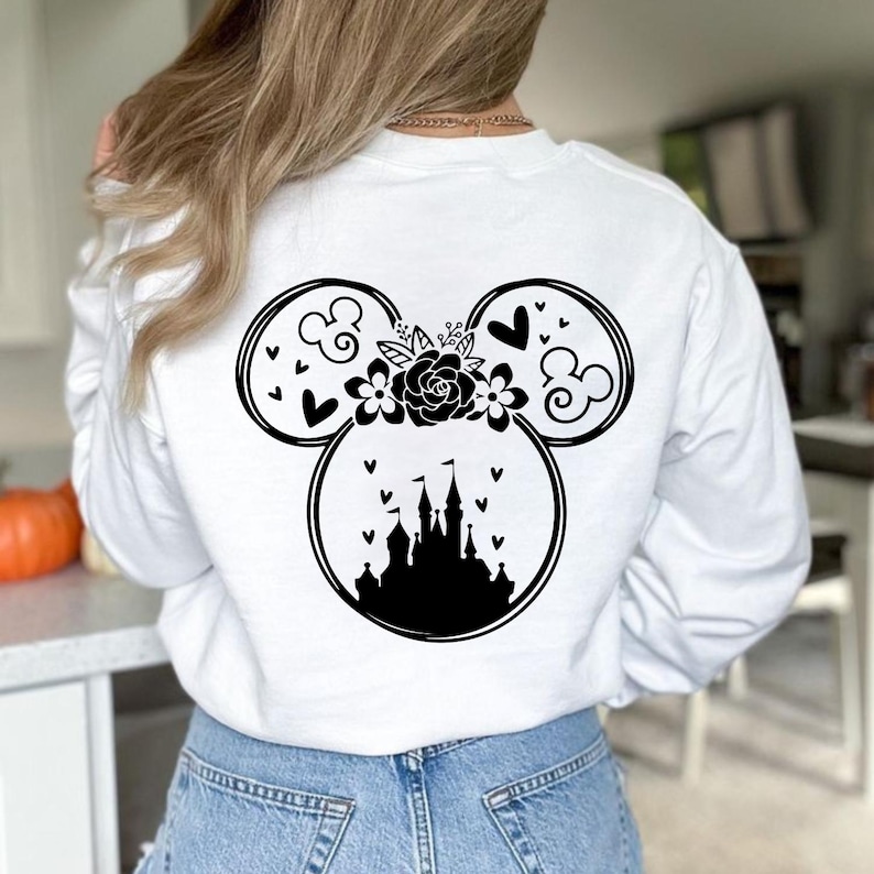 May include: White long-sleeve sweatshirt featuring a black Mickey Mouse design on the back. The design includes a castle, hearts, flowers, and swirls. The sweatshirt is made of a soft material.