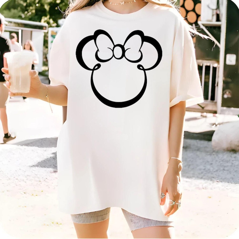 May include: Cream-colored t-shirt featuring a large black Minnie Mouse silhouette. The design includes a bow and the iconic mouse ears. The shirt is a relaxed fit, suitable for casual wear.