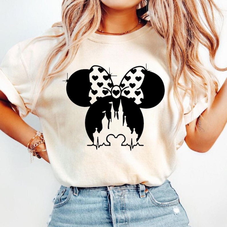 May include: Cream-colored t-shirt featuring a black graphic of a bow with heart patterns, Mickey Mouse ears, and a castle silhouette. A heartbeat line is below the castle. The shirt is a casual style.