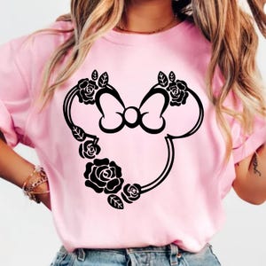 May include: A light pink t-shirt featuring a black Minnie Mouse silhouette design. The design includes a bow and is adorned with black roses and leaves. The shirt is made of a soft material.