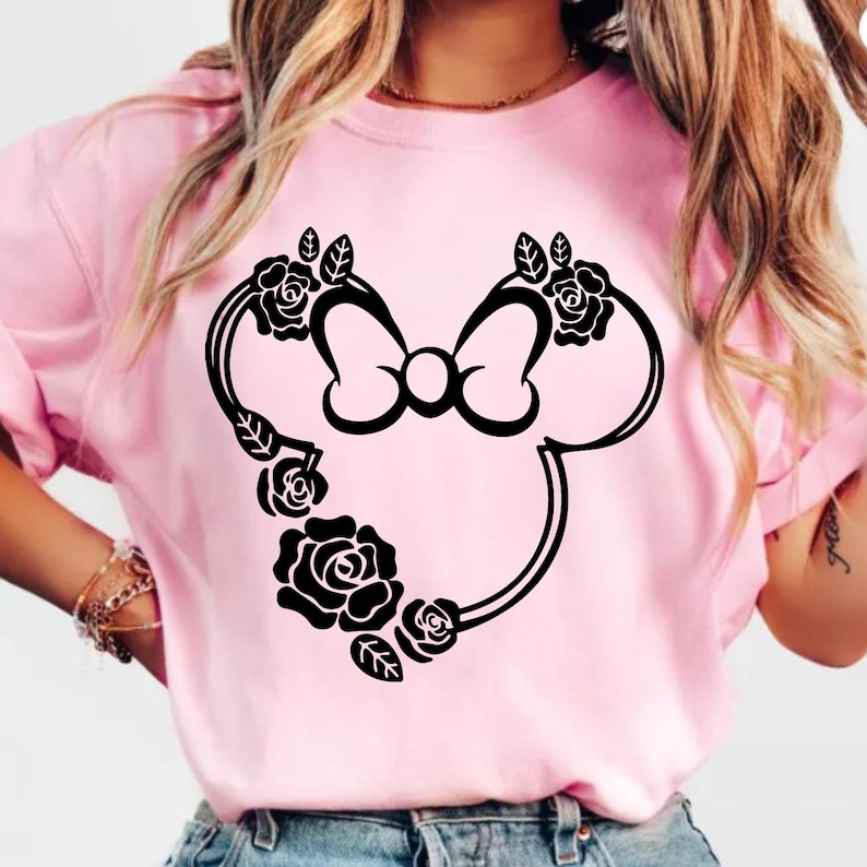 May include: A light pink t-shirt featuring a black Minnie Mouse silhouette design. The design includes a bow and is adorned with black roses and leaves. The shirt is made of a soft material.
