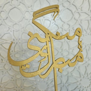 May include: A gold-colored wooden cutout of Arabic calligraphy. The calligraphy is in a decorative style and is mounted on a wooden stick.