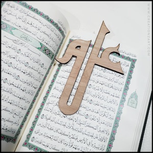 May include: A wooden bookmark with the Arabic word "Allah" carved into it. The bookmark is placed on an open Quran, a holy book in Islam.