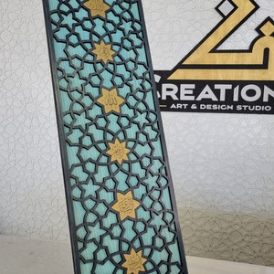 May include: A decorative wooden panel with a geometric pattern and gold accents. The panel has a teal background with a black overlay of interlocking shapes. Gold star-shaped elements with Arabic script are evenly spaced across the panel.