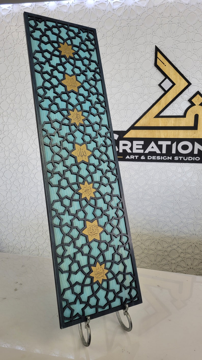 May include: A decorative wooden panel with a geometric pattern and gold accents. The panel has a teal background with a black overlay of interlocking shapes. Gold star-shaped elements with Arabic script are evenly spaced across the panel.