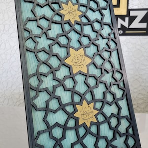May include: A decorative wooden panel featuring a geometric star pattern. The panel has a black frame and a light blue background. Gold star-shaped accents with Arabic calligraphy are featured. The panel is supported by a metal stand.