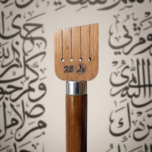 May include: A wooden weaving tool featuring a flat, rectangular head with four vertical slots and circular cutouts at the base of each slot. The number 25 and a small circular icon of a person kneeling are engraved on the light wood surface.