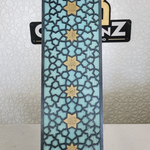 May include: A rectangular wooden panel with a geometric star pattern in black, over a light blue background. Gold-coloured Arabic calligraphy is featured within the stars. The panel is supported by two metal stands.