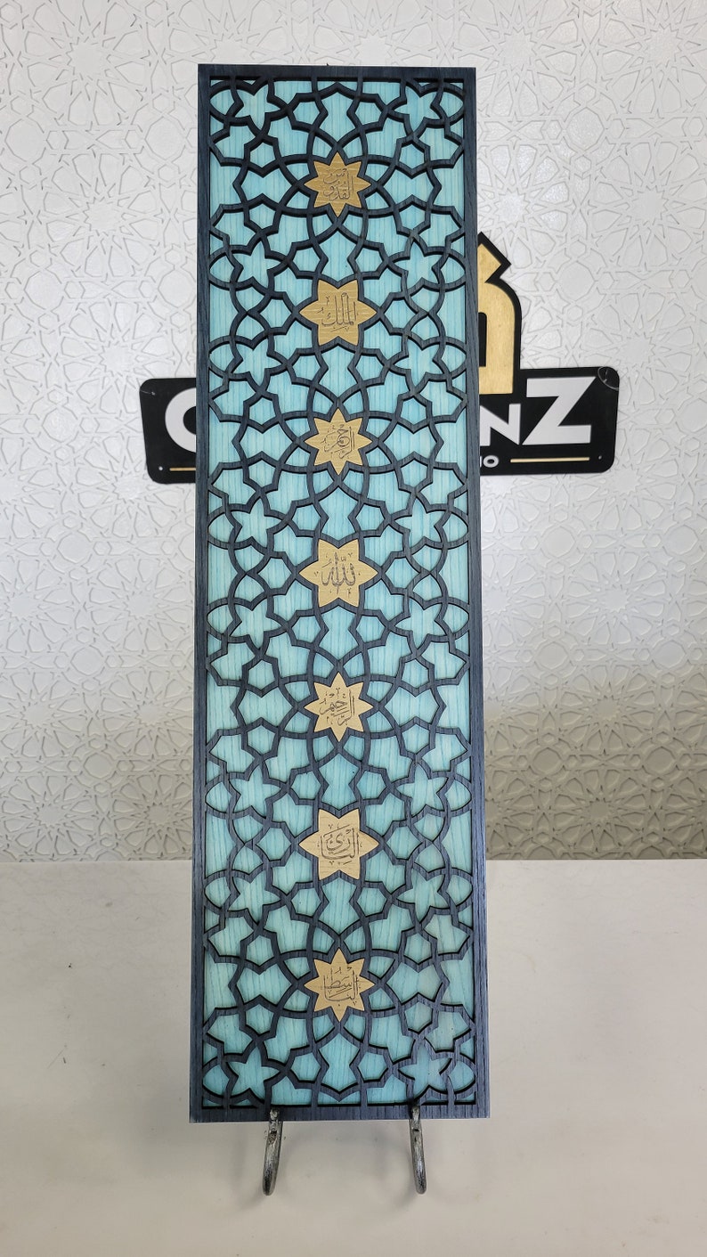May include: A rectangular wooden panel with a geometric star pattern in black, over a light blue background. Gold-coloured Arabic calligraphy is featured within the stars. The panel is supported by two metal stands.