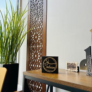 May include: A square black wooden sign featuring a gold circular geometric design with the word Salaam written in both Arabic script and English text. The sign is positioned on a wooden surface next to a small rectangular frame with floral details.