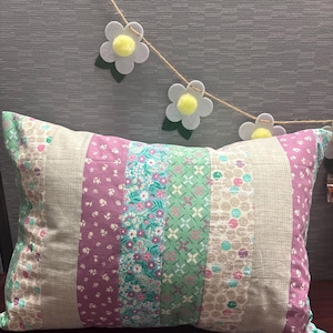 May include: A rectangular patchwork pillow with vertical stripes in various floral and patterned fabrics. Colors include pink, green, blue, and beige. A daisy garland hangs above the pillow.