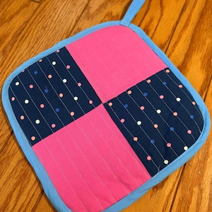 May include: A square hot pad with a blue border and a patchwork design. The center is pink with two squares of navy blue fabric with white and pink polka dots.