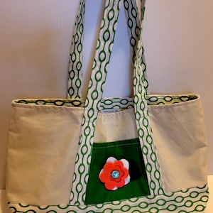 May include: A green and white patterned tote bag with a green pocket and a red flower with a blue button in the center. The bag has a label that says "handmade by"