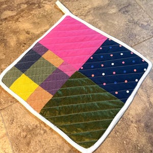 May include: A quilted pot holder with a patchwork design. The pot holder features a pink square, a navy blue square with white and pink polka dots, a green square, and a plaid square with yellow, orange, and purple. It has a white border and a hanging loop.