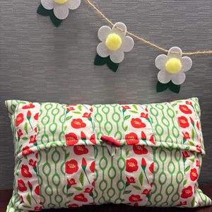 May include: A rectangular pillow with a white background and a pattern of red flowers and green stems. The pillow has a red button closure. A garland of felt flowers with yellow centers hangs above the pillow.