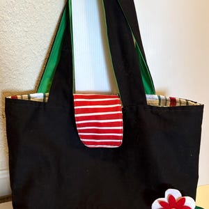 May include: Black canvas tote bag with green straps, a red and white striped pocket, and a white felt flower with a red center.