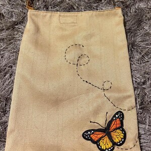 May include: Beige canvas crossbody bag with a butterfly embroidery in orange, yellow, and black. The bag has a herringbone pattern and brown leather straps with metal rings.