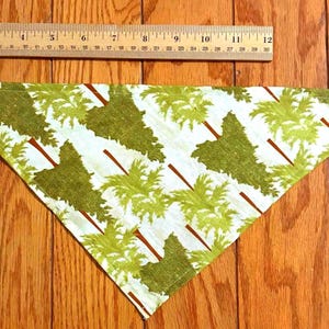 May include: Triangular pet bandana with a white background and a repeating pattern of green palm trees with brown trunks. A wooden ruler is placed above the bandana, measuring up to 12 inches.