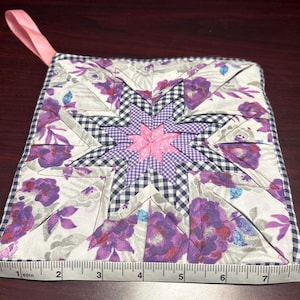 May include: A square quilted potholder featuring a central star pattern in pink and purple gingham, surrounded by a white fabric with purple floral prints. A pink fabric loop is attached to one corner. A measuring tape shows a width of 8 inches.