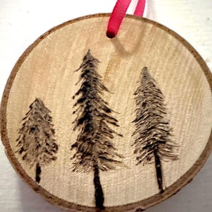 May include: A round wooden ornament with a natural wood grain finish. Three stylized evergreen trees are burned into the wood, with a dark brown color. A bright pink ribbon is attached for hanging. The ornament is a rustic, handmade item.