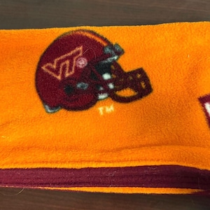 May include: Orange fleece blanket featuring a maroon football helmet with the letters "VT" and the word "VIRGINIA". The blanket has a maroon border.