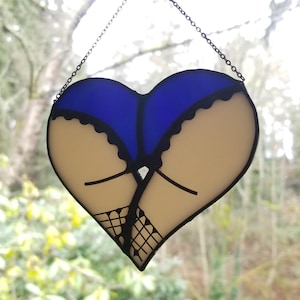 May include: A heart-shaped stained glass piece depicting a stylized rear view. The design features a blue top and tan bottom, with black accents and fishnet stockings. A chain is attached for hanging.