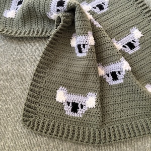 May include: A handmade, olive green crochet blanket featuring repeating koala bear designs. The koalas are depicted with white, gray, and black details. The blanket is folded, showcasing the pattern and texture of the yarn.