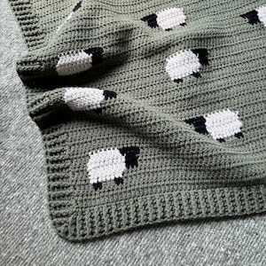 Könnte beinhalten: Handgefertigte Häkeldecke in einem sanften Grünton, verziert mit sich wiederholenden weißen Schafen mit schwarzen Gesichtern und Beinen. Die Decke hat ein strukturiertes Stichmuster und einen fertigen Rand, perfekt für ein Kinderzimmer oder die Inneneinrichtung.