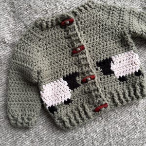 May include: A handmade, olive green crochet baby cardigan with three red buttons. The cardigan features two sheep appliqués with white and black wool. The garment is displayed on a gray, textured surface.