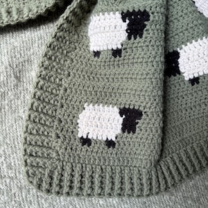 May include: A crocheted blanket in shades of green, featuring a repeating pattern of white sheep with black faces and legs. The blanket has a textured appearance and a ribbed border, perfect for a nursery or as a decorative throw.