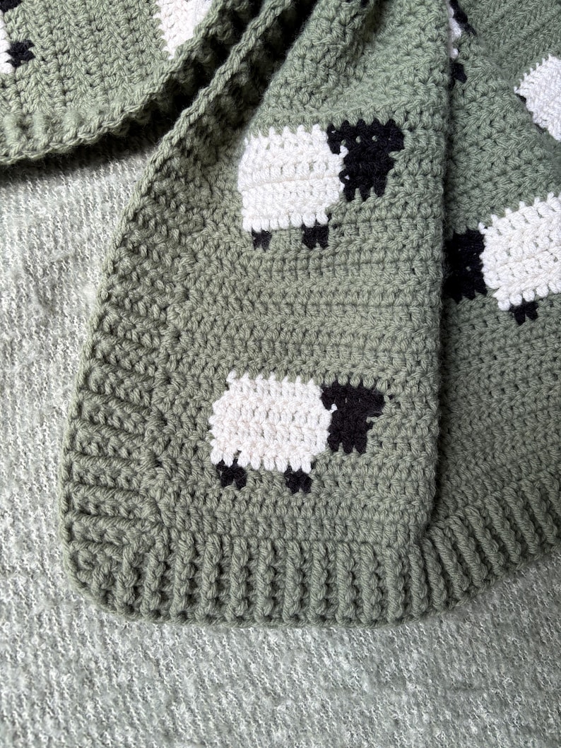 May include: A crocheted blanket in shades of green, featuring a repeating pattern of white sheep with black faces and legs. The blanket has a textured appearance and a ribbed border, perfect for a nursery or as a decorative throw.