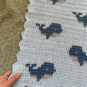 以下が含まれることがあります： 水色のスカラップエッジのかぎ針編みブランケット。ブランケットには、青とタンの色合いのクジラの繰り返し模様があります。ブランケットは手作りで、柔らかく居心地の良いように見えます。