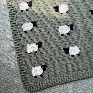 May include: Crocheted blanket in shades of green, featuring a pattern of white sheep with black faces and legs. The blanket has a textured appearance and a ribbed border. The sheep are evenly spaced across the surface.
