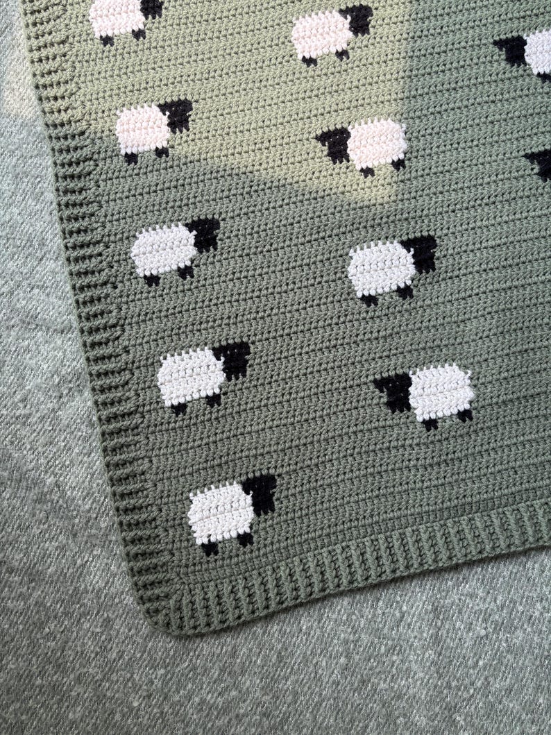 May include: Crocheted blanket in shades of green, featuring a pattern of white sheep with black faces and legs. The blanket has a textured appearance and a ribbed border. The sheep are evenly spaced across the surface.