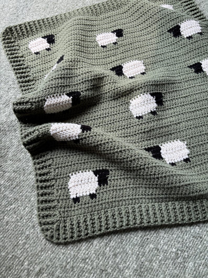 May include: A handmade crochet blanket in a soft green hue, adorned with repeating white sheep with black faces and legs. The blanket features a textured stitch pattern and a finished edge, perfect for a nursery or home decor.